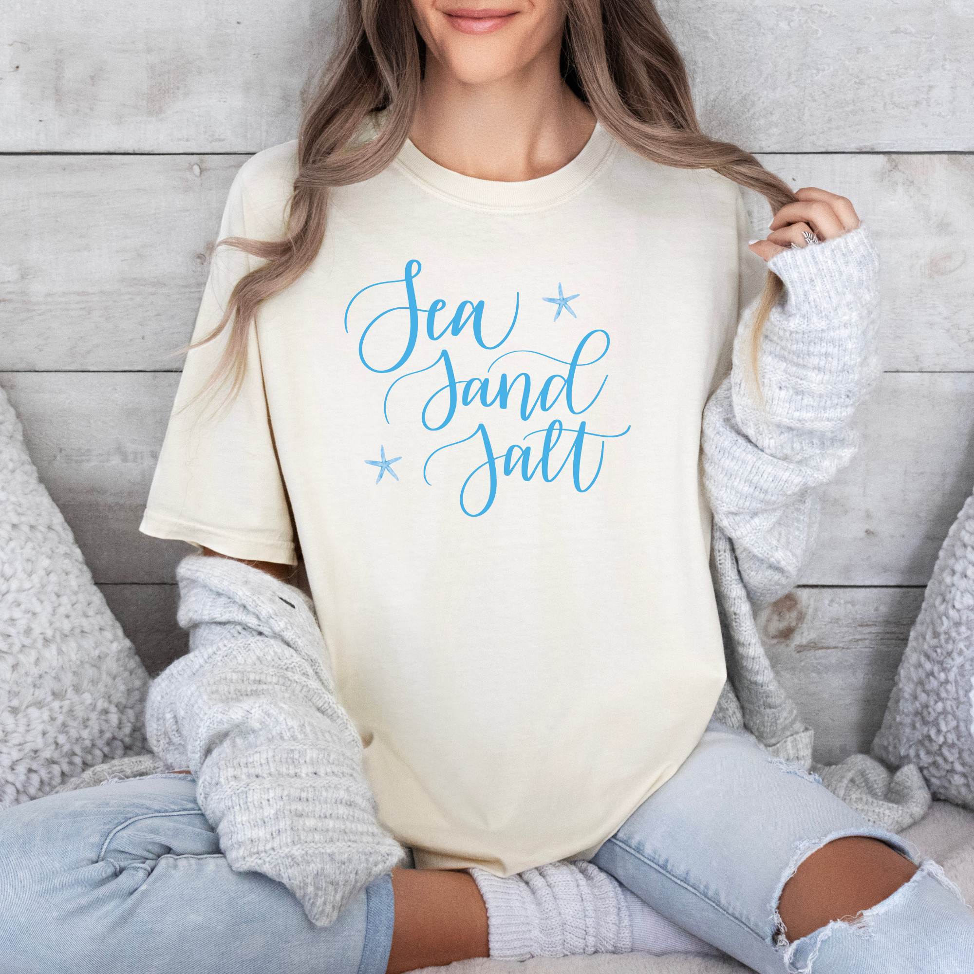 Person wearing a cream-colored sweatshirt with 'Sea Sand Salt' text in blue, sitting on a wooden floor.