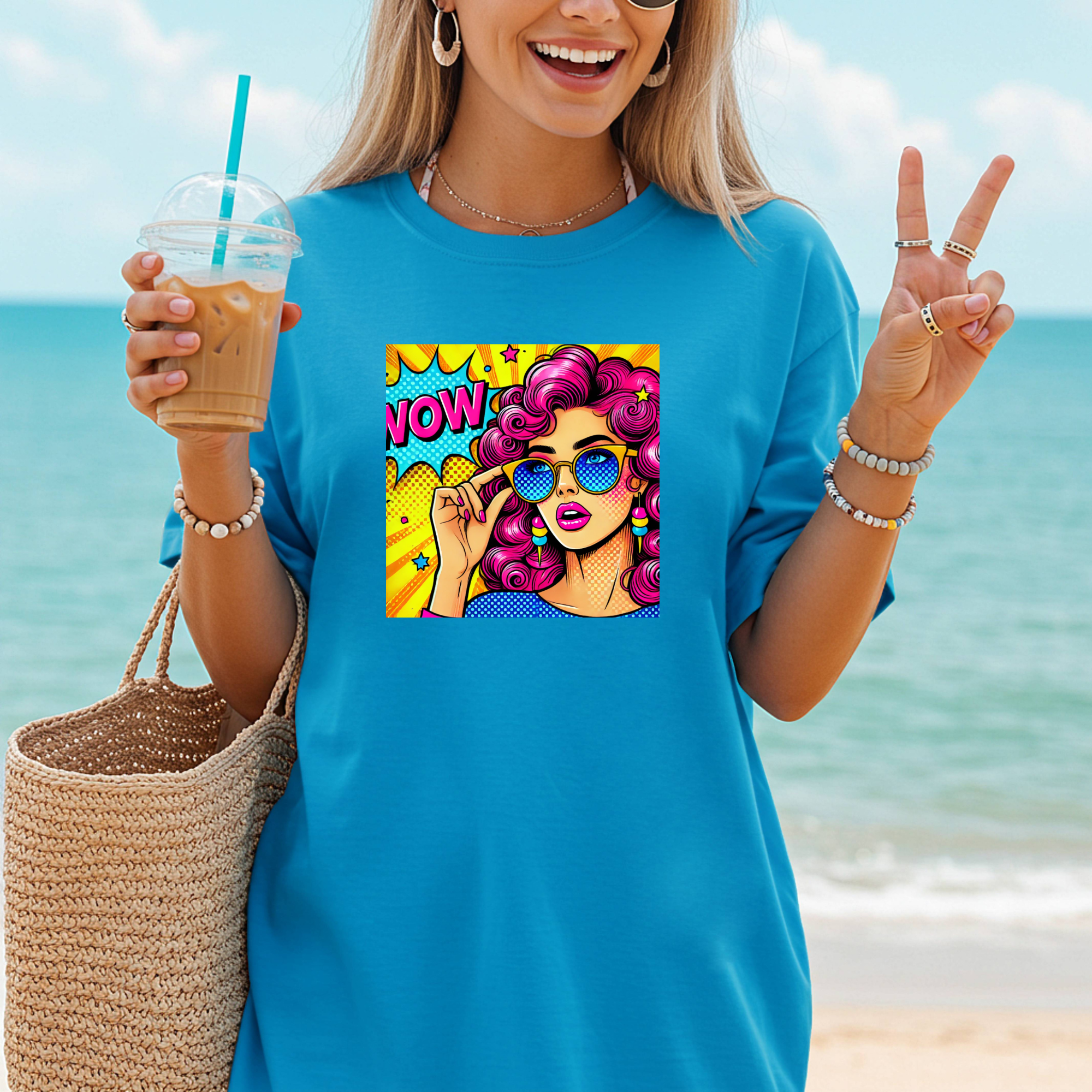 Woman on a beach wearing a blue t-shirt with a colorful graphic design, holding a drink.