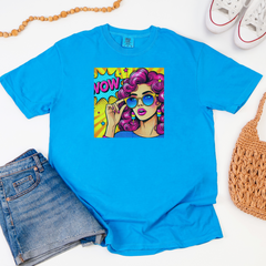 Blue t-shirt with a colorful graphic of a woman with sunglasses and 'NOW' text, surrounded by jeans, sneakers, and a bag on a white background.