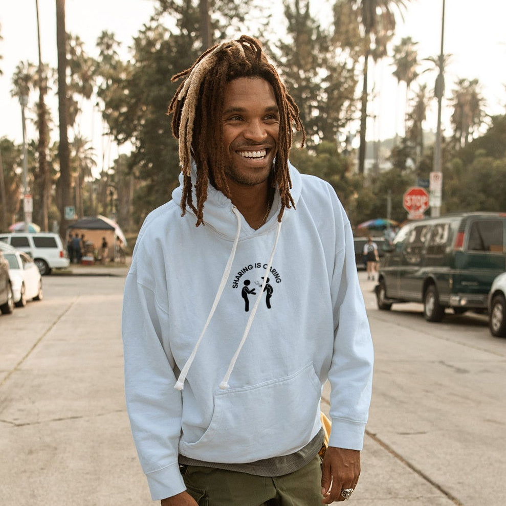 Man in a light blue hoodie and green pants standing on a skateboard in an urban setting with palm trees.