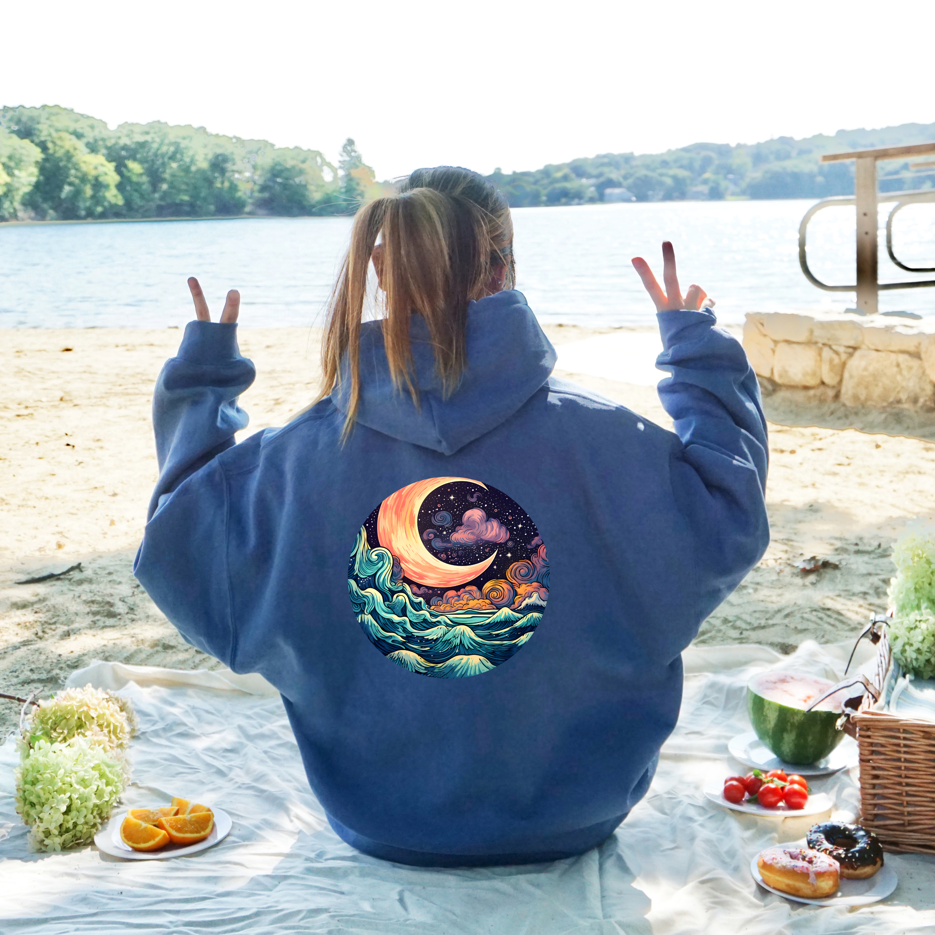 Person wearing a blue hoodie with a celestial design by a lakeside picnic.