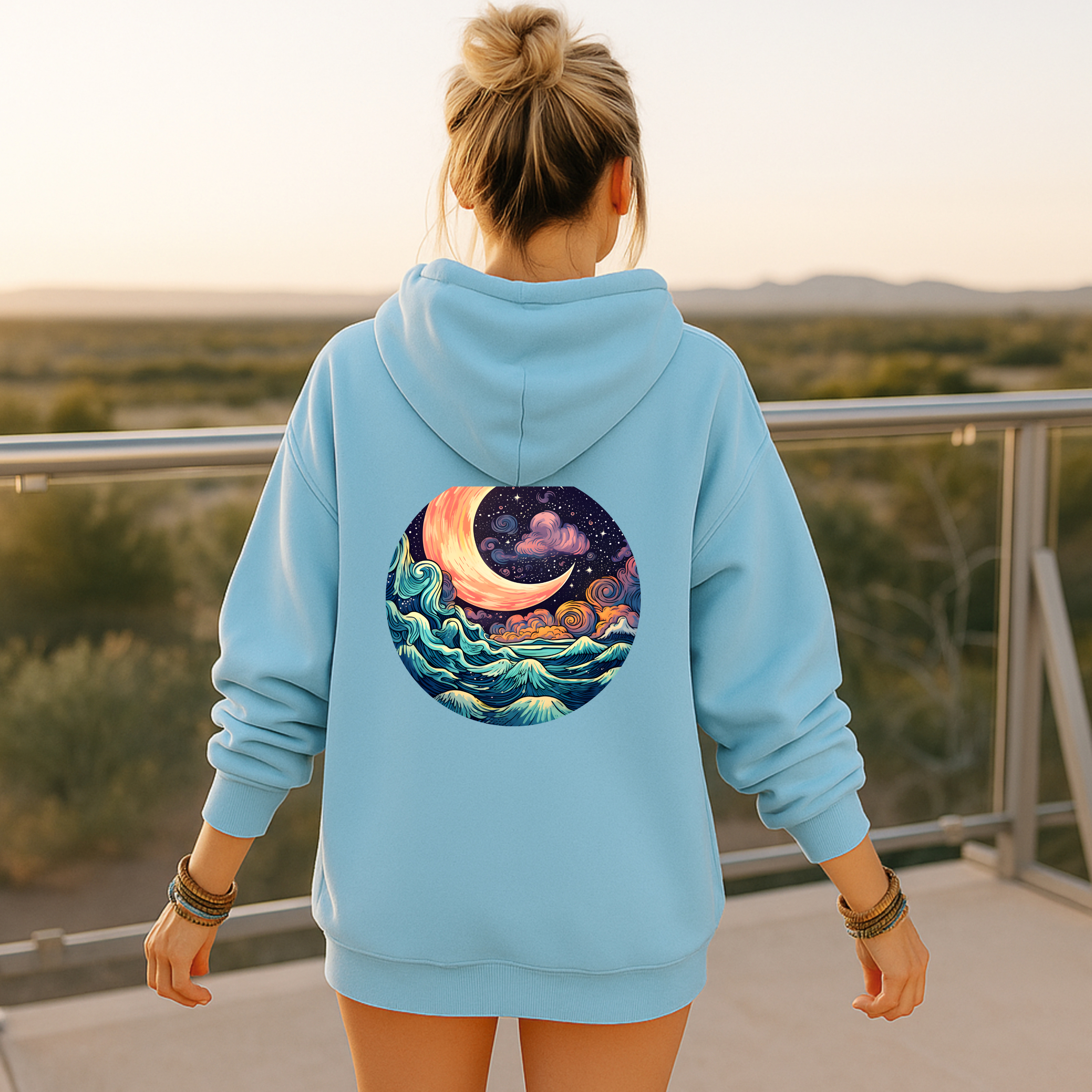 Person wearing a light blue hoodie with a celestial design on the back, standing on a balcony with a scenic view.
