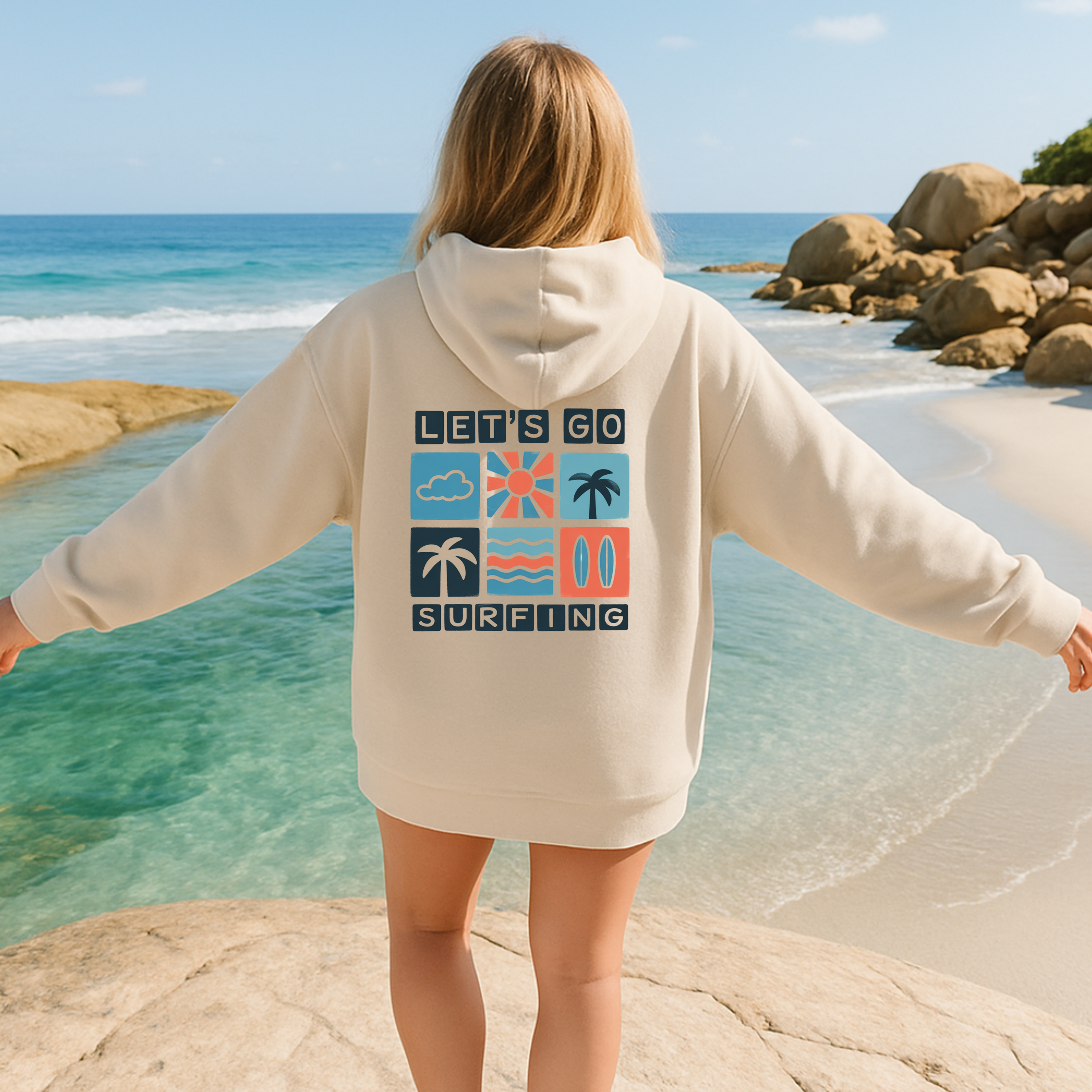 Person wearing a hoodie with 'Let's Go Surfing' text and graphics on a beach.