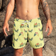 Person wearing green parrot-patterned swim shorts on a rocky beach.