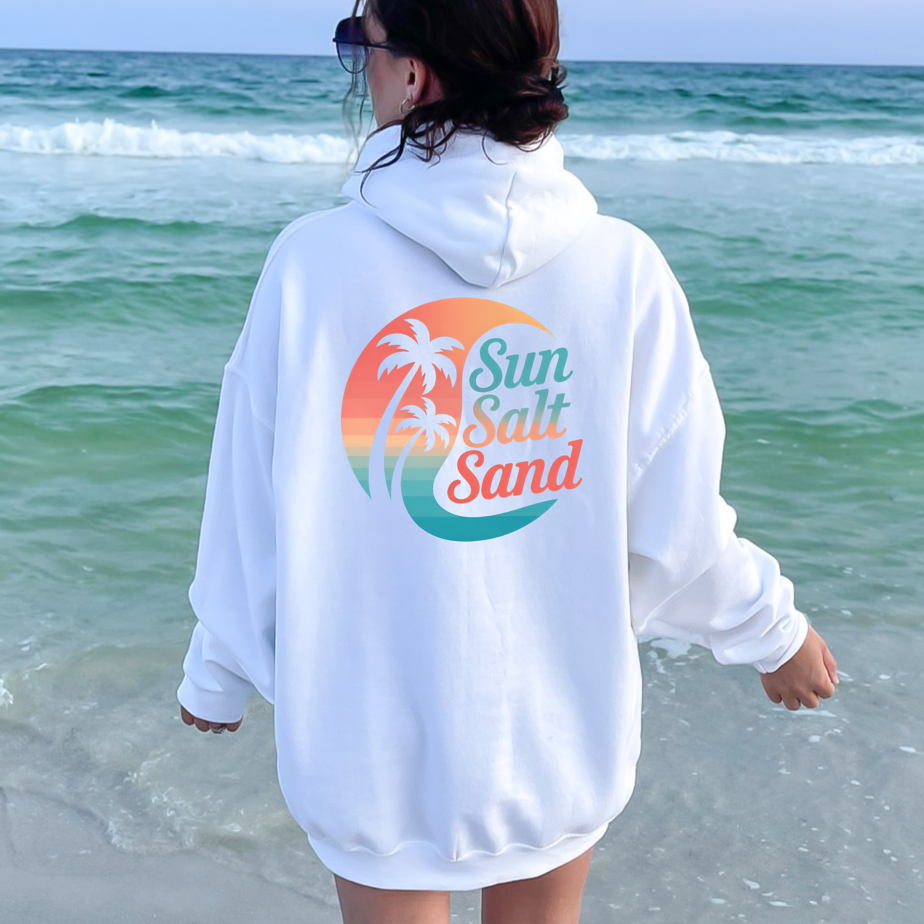 Person wearing a white hoodie with 'Sun Salt Sand' design on a beach