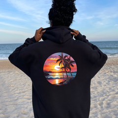 Person wearing a black hoodie with a tropical sunset design on a beach