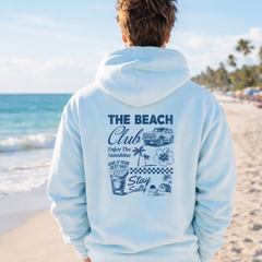 Person wearing a light blue hoodie with 'The Beach Club' design on a beach.
