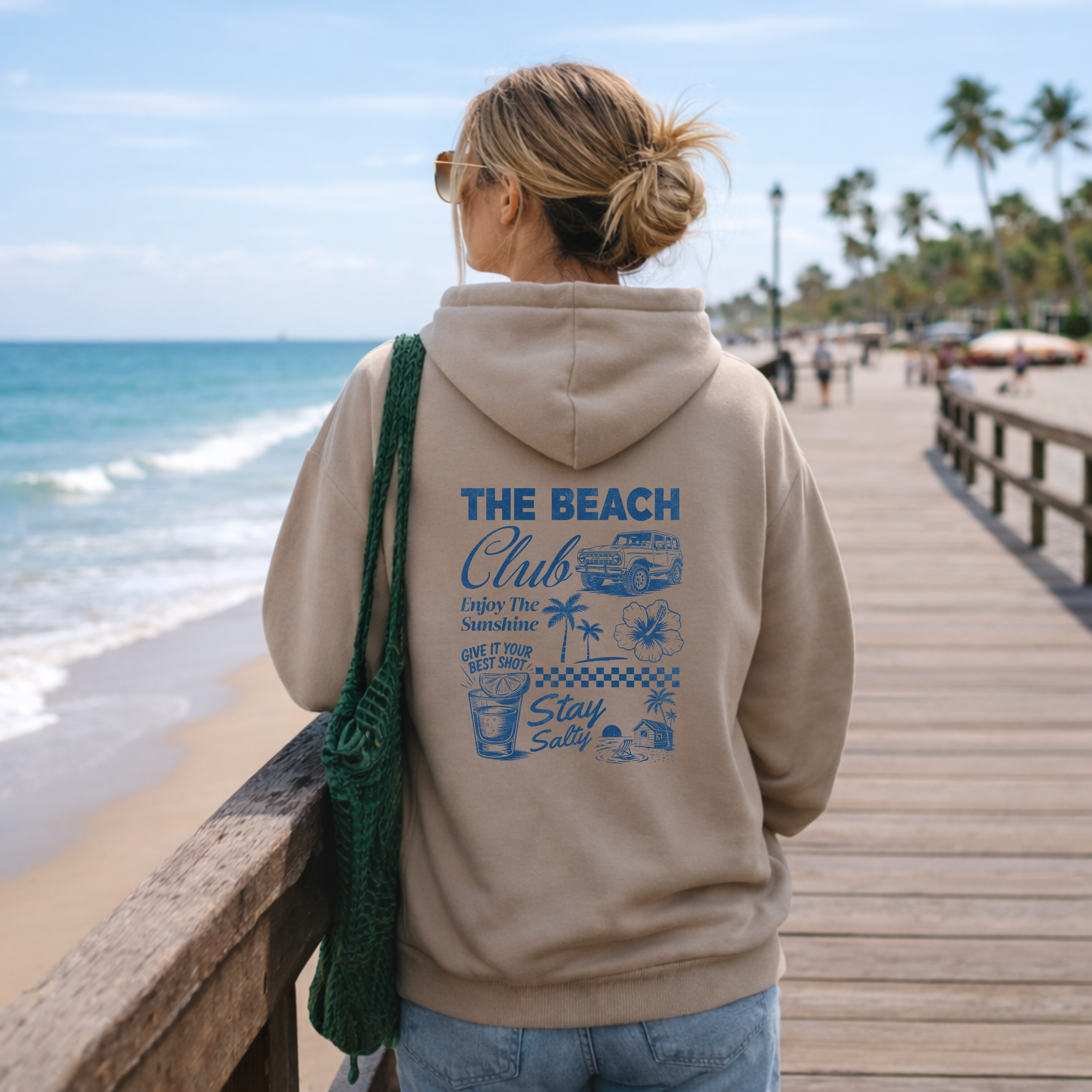 Person wearing a hoodie with 'The Beach Club' design on a beach boardwalk.