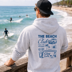 Person wearing a hoodie with 'The Beach Club' design by the ocean.