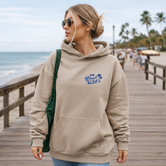 Woman wearing a beige hoodie with 'Beach Club' logo on a wooden boardwalk by the beach.