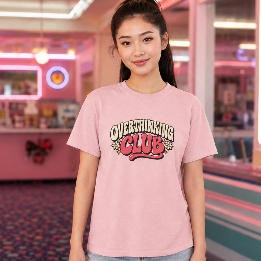 A young woman stands in a retro-themed arcade or diner interior wearing a light pink graphic t-shirt that reads 'OVERTHINKING CLUB' and light-wash jeans.