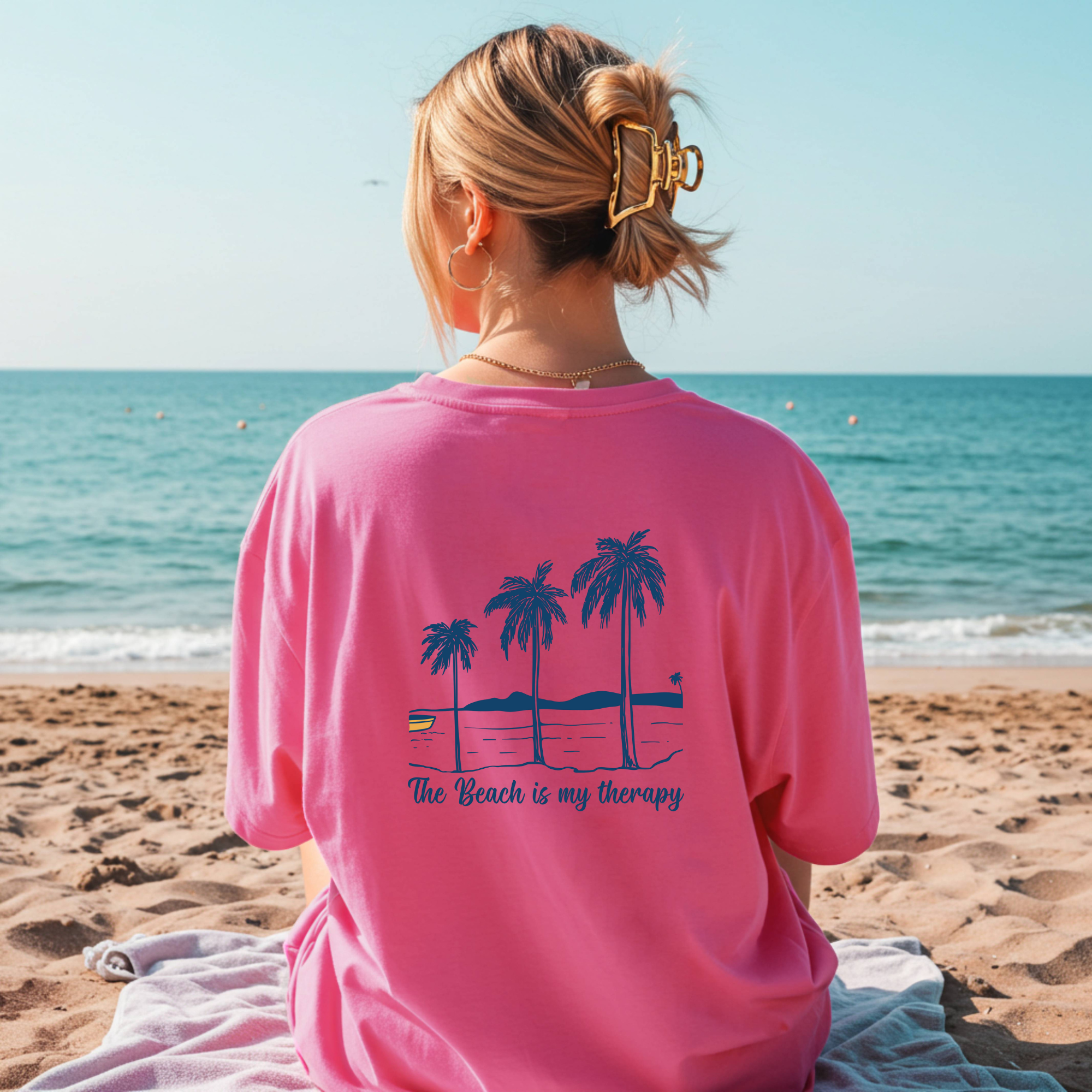 Person wearing a pink shirt with palm tree design and 'The Beach is my Therapy' text on a beach.
