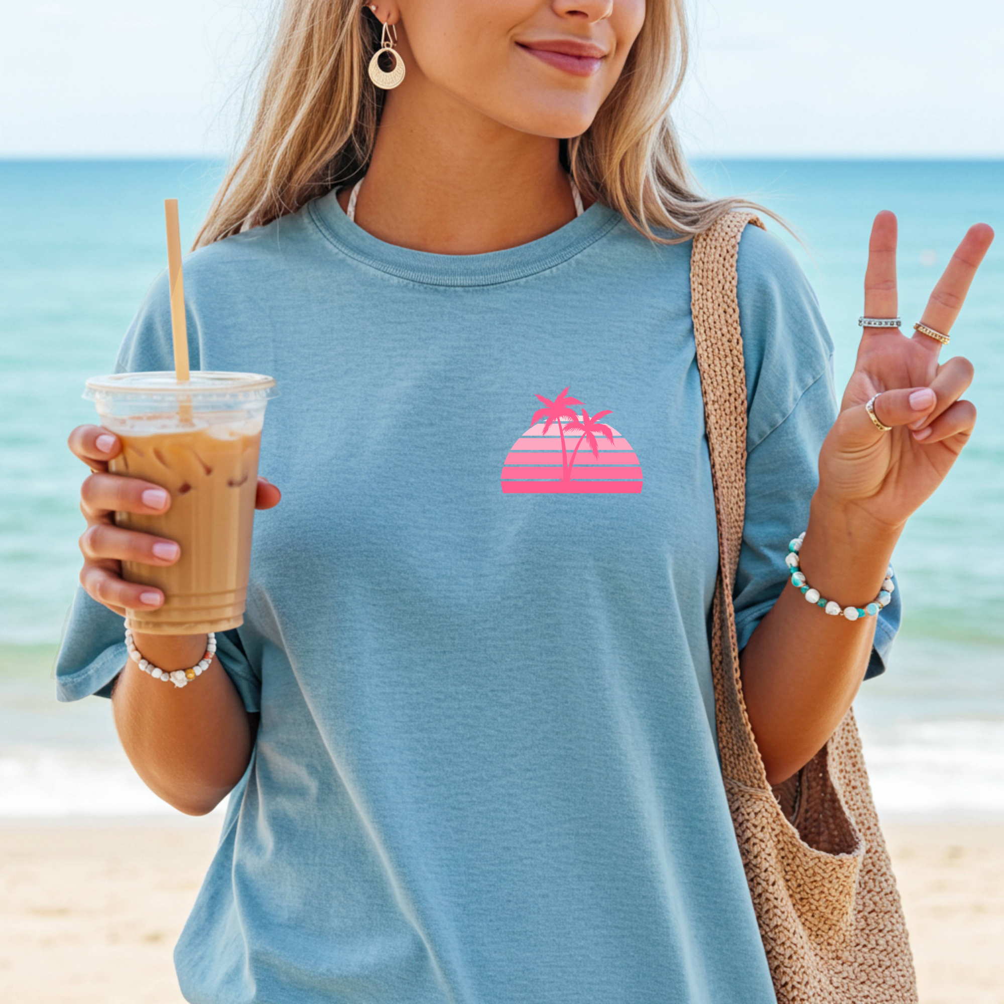 Woman on a beach holding a drink and making a peace sign, wearing a blue t-shirt with a pink palm tree design.