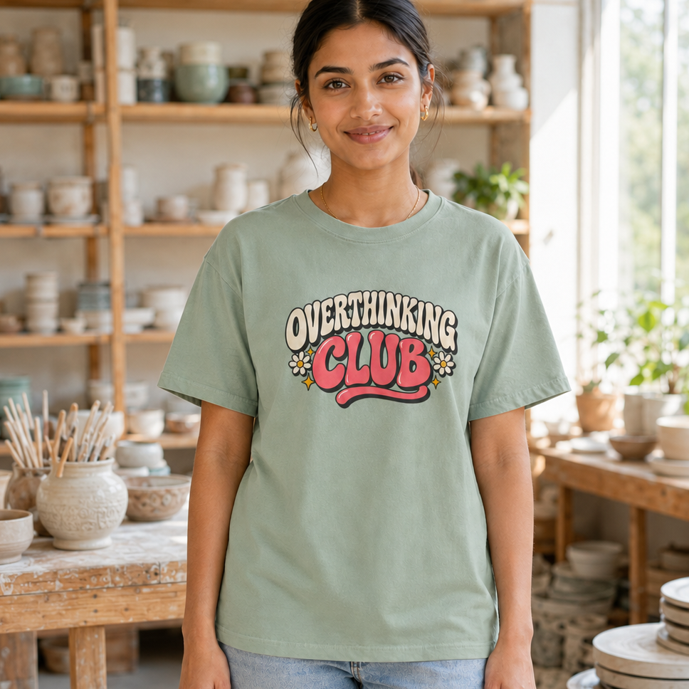 A woman wearing a sage green graphic t-shirt that reads 'OVERTHINKING CLUB' in a retro red and cream design, standing in a pottery studio filled with ceramic shelves and tables.