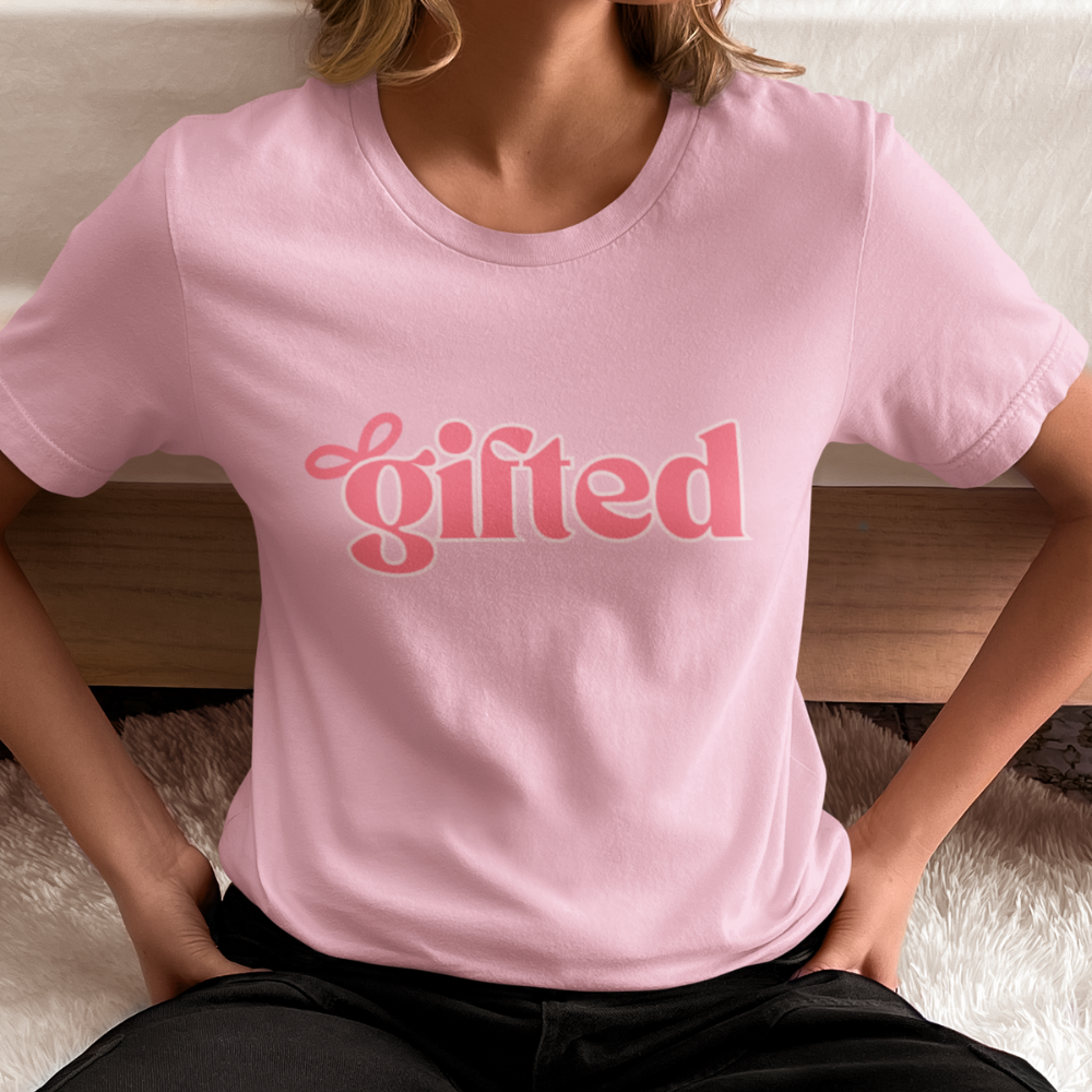 Person wearing a pink t-shirt with 'gifted' printed on it, sitting on a textured surface.