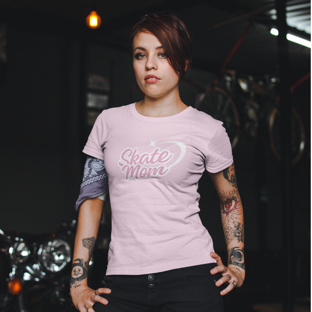Woman wearing a pink 'Skate Mom' t-shirt with tattoos on her arms, standing in a dark indoor setting.