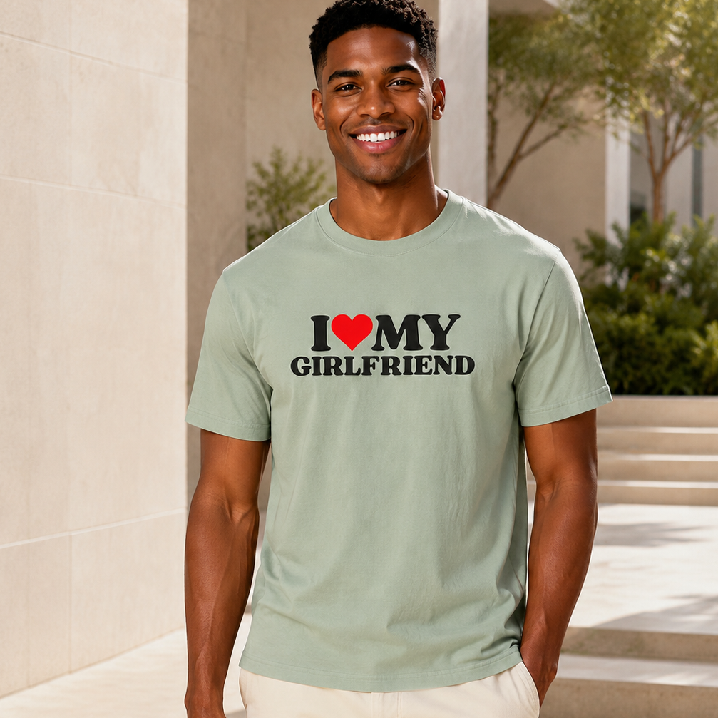 A man wearing a sage green t-shirt with a graphic that reads 'I [red heart] MY GIRLFRIEND' in black and red text, paired with off-white pants, standing outdoors on stone steps.