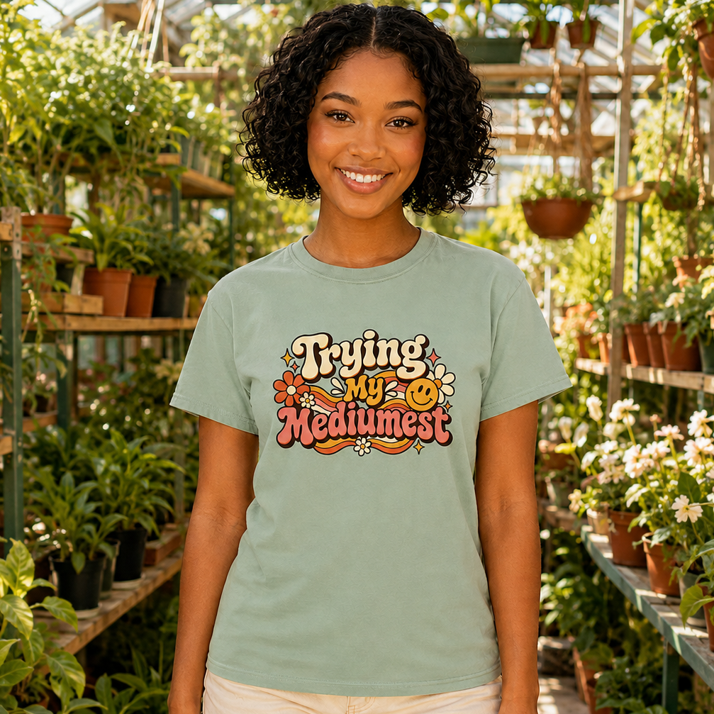 A smiling woman stands in a plant-filled greenhouse wearing a sage green graphic T-shirt that reads 'Trying My Life Medumest' with a small smiley face and flowers, paired with light beige shorts.