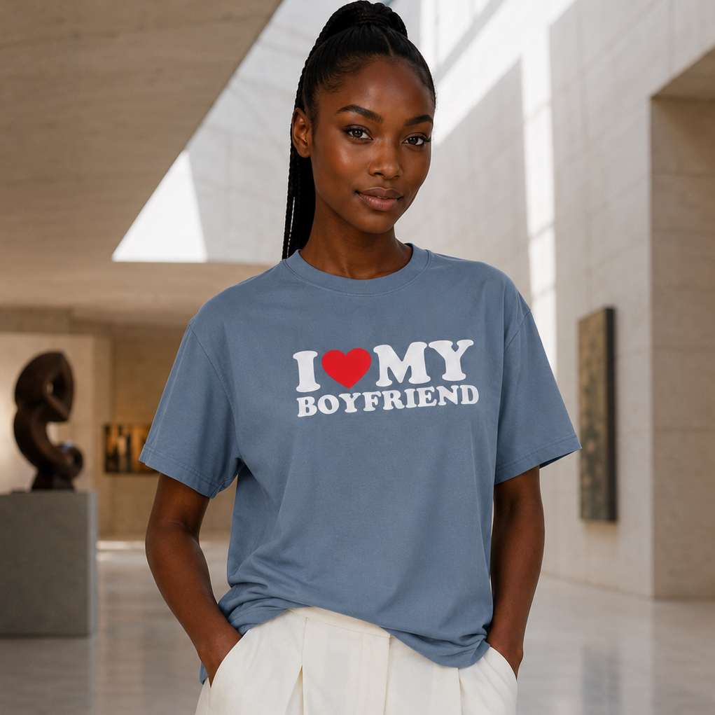 A woman wearing a slate blue graphic t-shirt that reads 'I [red heart] MY BOYFRIEND' in white and red, paired with white pants, standing in a modern art gallery.