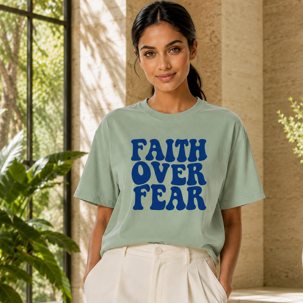 A woman stands outdoors by a pool wearing a light sage green graphic T-shirt with the words 'FAITH OVER FEAR' printed in bold blue letters, paired with white pants.