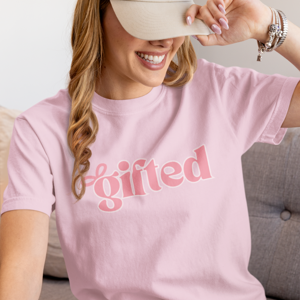 Woman wearing a pink t-shirt with 'gifted' text, smiling and holding a beige visor.