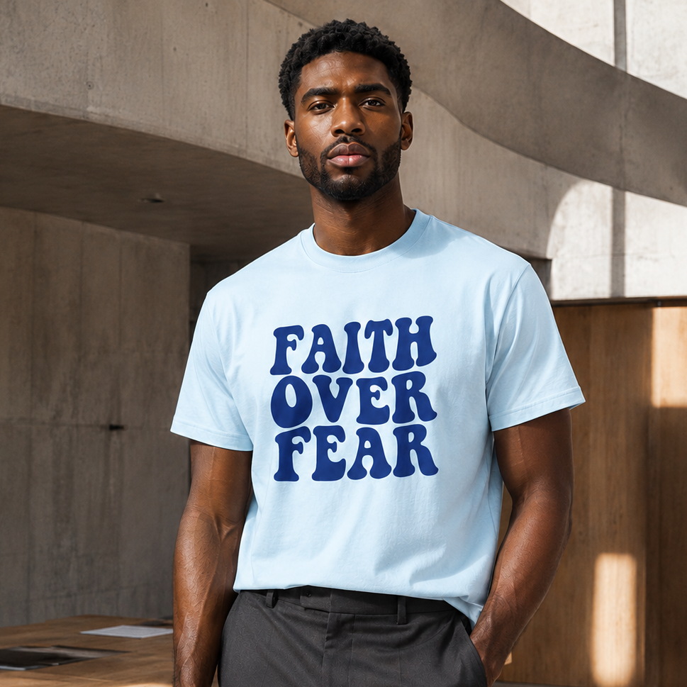 A man wearing a light blue crewneck t-shirt with the words 'FAITH OVER FEAR' printed in dark blue block letters on the front, paired with dark gray trousers, standing in a modern concrete and wood interior.