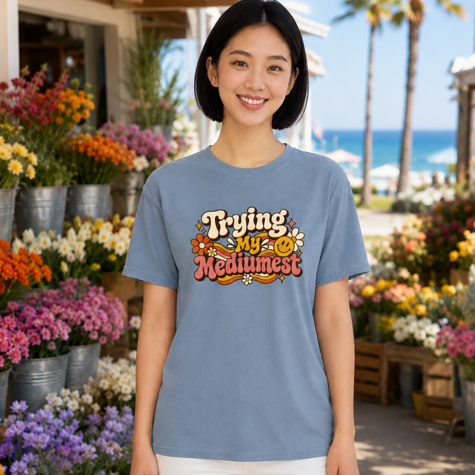 A woman stands outdoors in a sunny flower market wearing a light blue graphic T-shirt with the phrase 'Trying My Medimost' in a colorful retro design, paired with white pants.