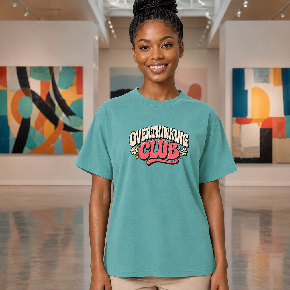 A woman stands in an art gallery wearing a teal graphic t-shirt that reads 'OVERTHINKING CLUB' in white, pink, and red lettering, paired with beige pants.