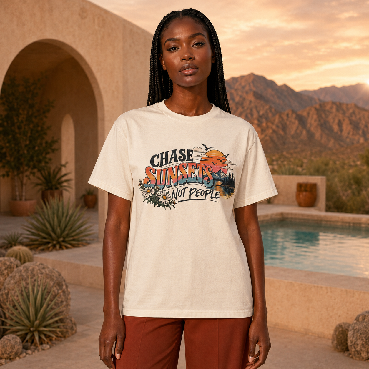A woman stands outdoors in front of a desert pool and mountains, wearing a cream graphic T-shirt with the text 'Chase Sunsets Not People' and a sunset illustration, paired with rust-colored pants.