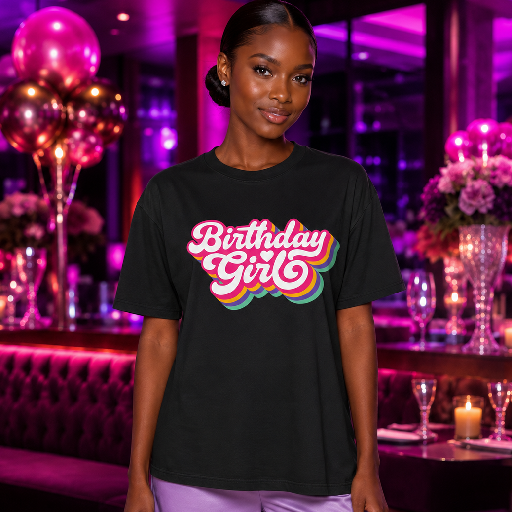 A woman stands in a dimly lit, purple-themed party venue wearing a black graphic t-shirt with 'Birthday Girl' in a pink, orange, and teal retro bubble font, paired with lavender pants.