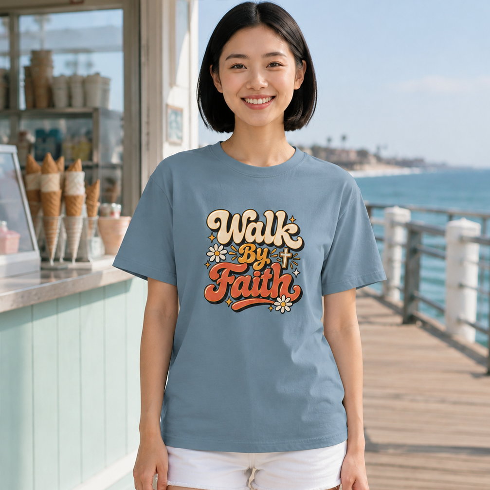 A woman stands on a wooden boardwalk by the ocean wearing a light blue graphic t-shirt with 'Walk By Faith' printed on the front in orange, yellow, and white lettering, paired with white shorts.