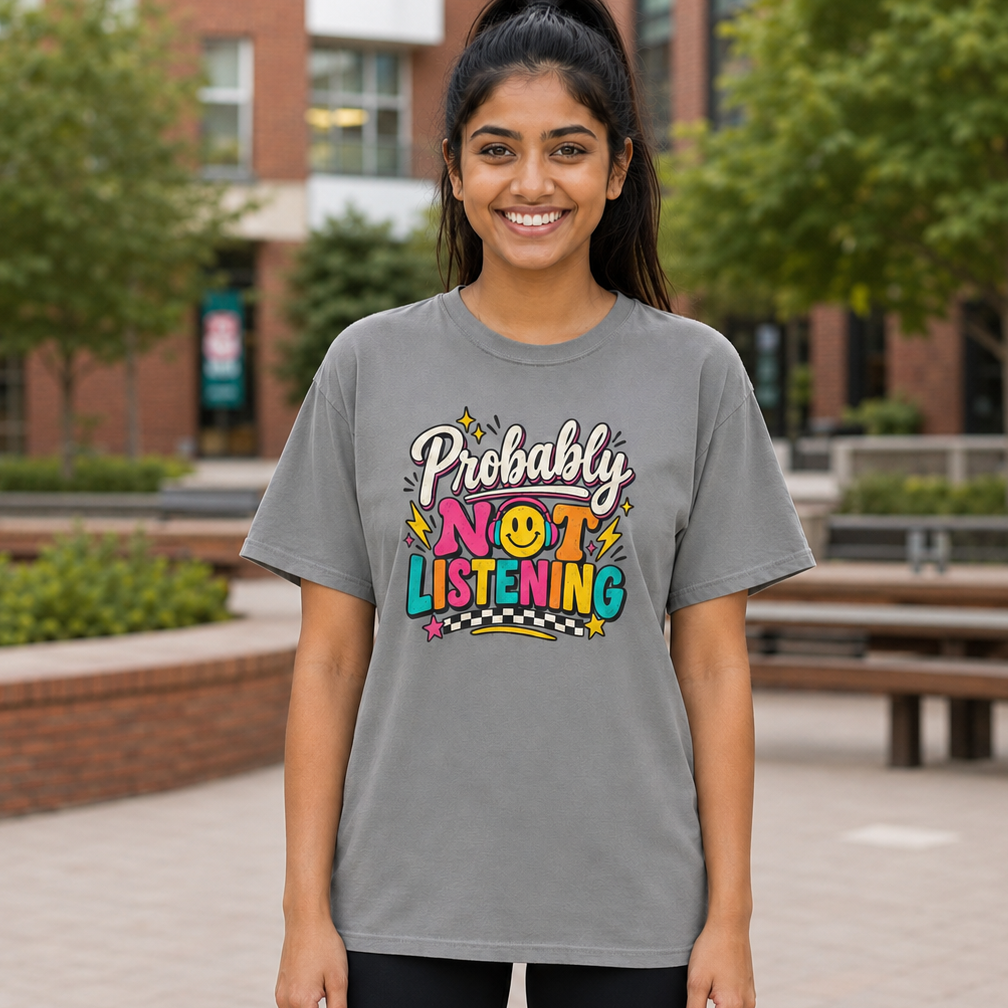 A woman standing outdoors in front of a brick building and trees, wearing a gray graphic T-shirt with the phrase 'Probably Not Listening' in colorful lettering and black shorts.