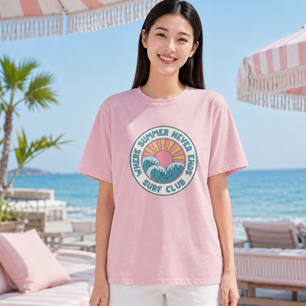 A woman stands on a beach patio wearing an oversized light pink t-shirt with a colorful circular 'Night Summer Never Sleep Surf Club' graphic on the chest, paired with white shorts.