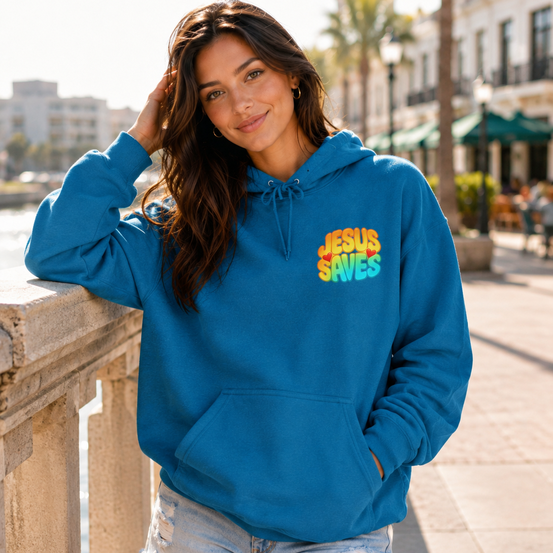 A woman wearing a bright blue hoodie with a colorful 'JESUS SAVES' graphic on the chest, posing outdoors on a sunny day next to a stone railing.
