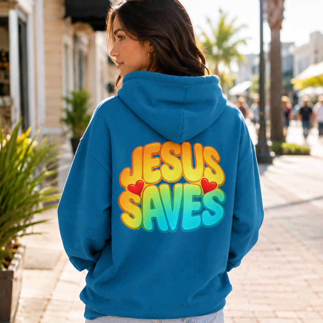 A woman standing outdoors on a sunny sidewalk, shown from the back wearing a bright blue hoodie with a large colorful graphic on the back that reads 'JESUS SAVES' in yellow, orange, and blue gradient letters with small red hearts.
