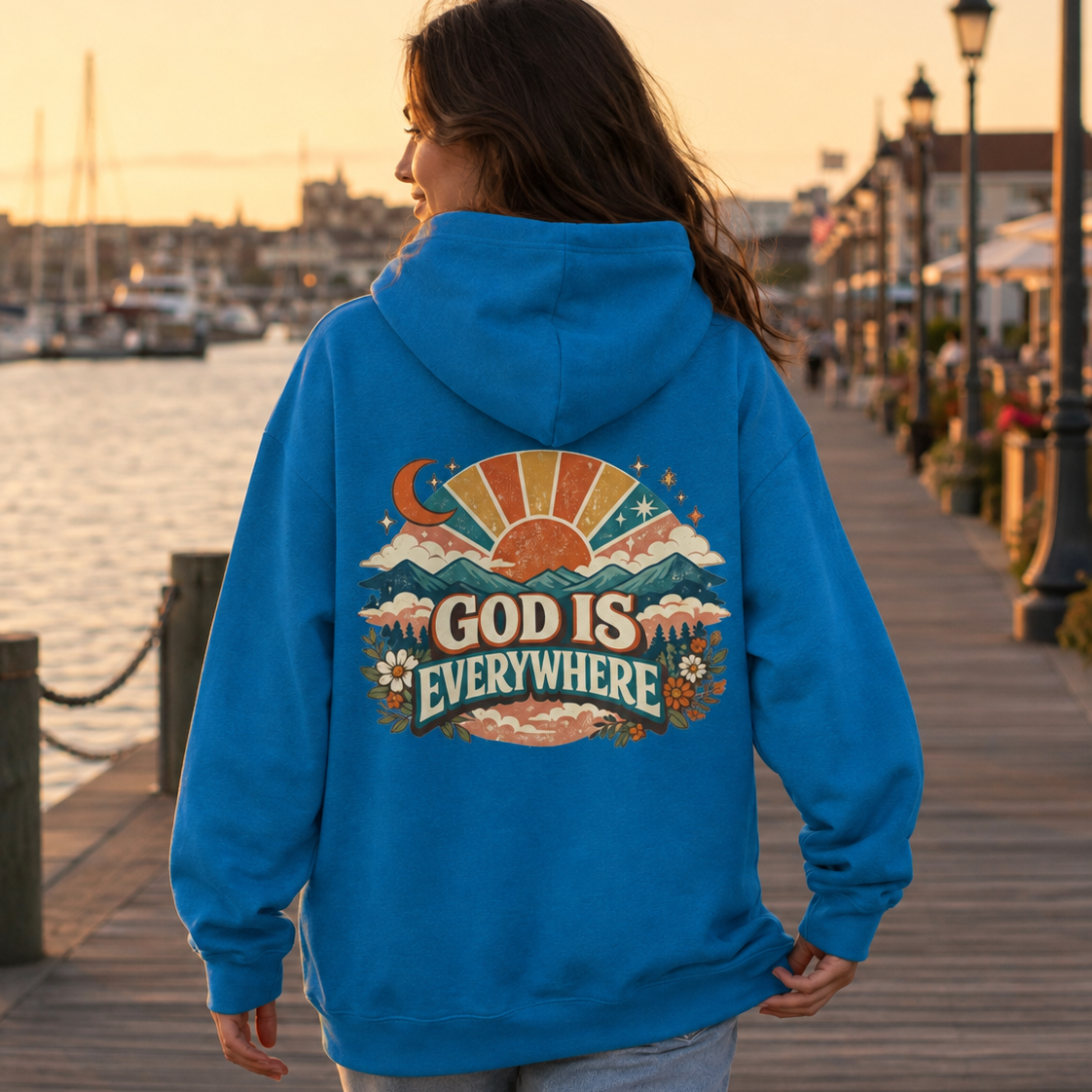 A woman viewed from behind wearing a bright blue hoodie with a large retro-style multicolor graphic on the back that reads “GOD IS EVERYWHERE,” standing on a wooden boardwalk by the water at sunset.