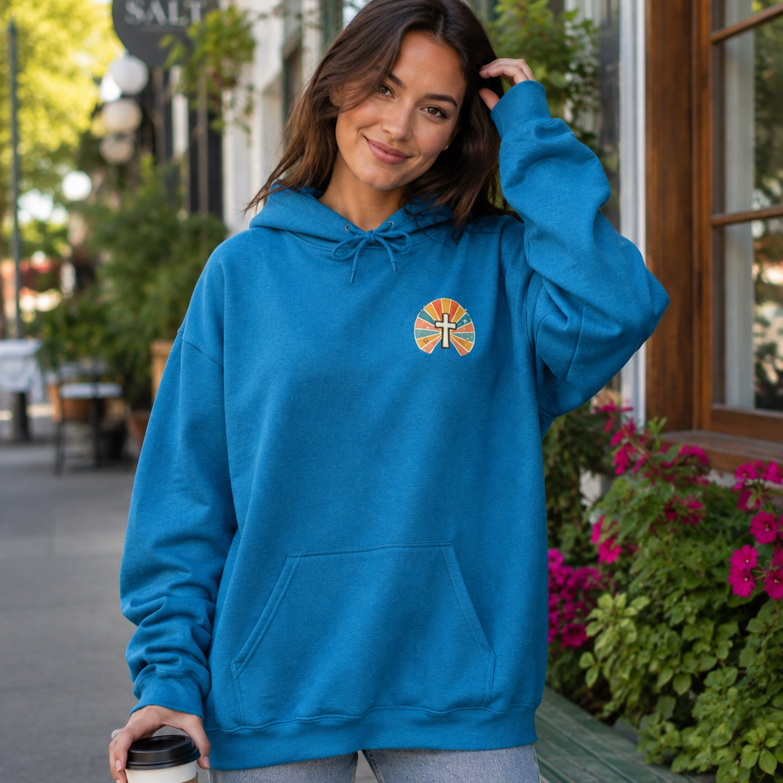 A woman wearing a bright blue pullover hoodie with a small circular orange and yellow graphic on the left chest, standing on a city sidewalk and holding a takeaway coffee cup.