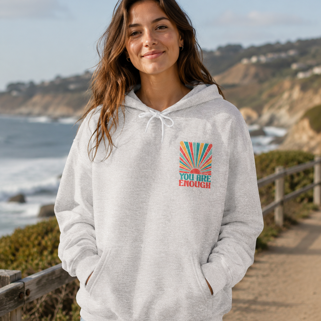 A woman wearing a light gray heather pullover hoodie with a colorful graphic on the left chest that reads 'YOU ARE ENOUGH' with a rainbow sunburst, standing on a beach boardwalk with the ocean and cliffs in the background.