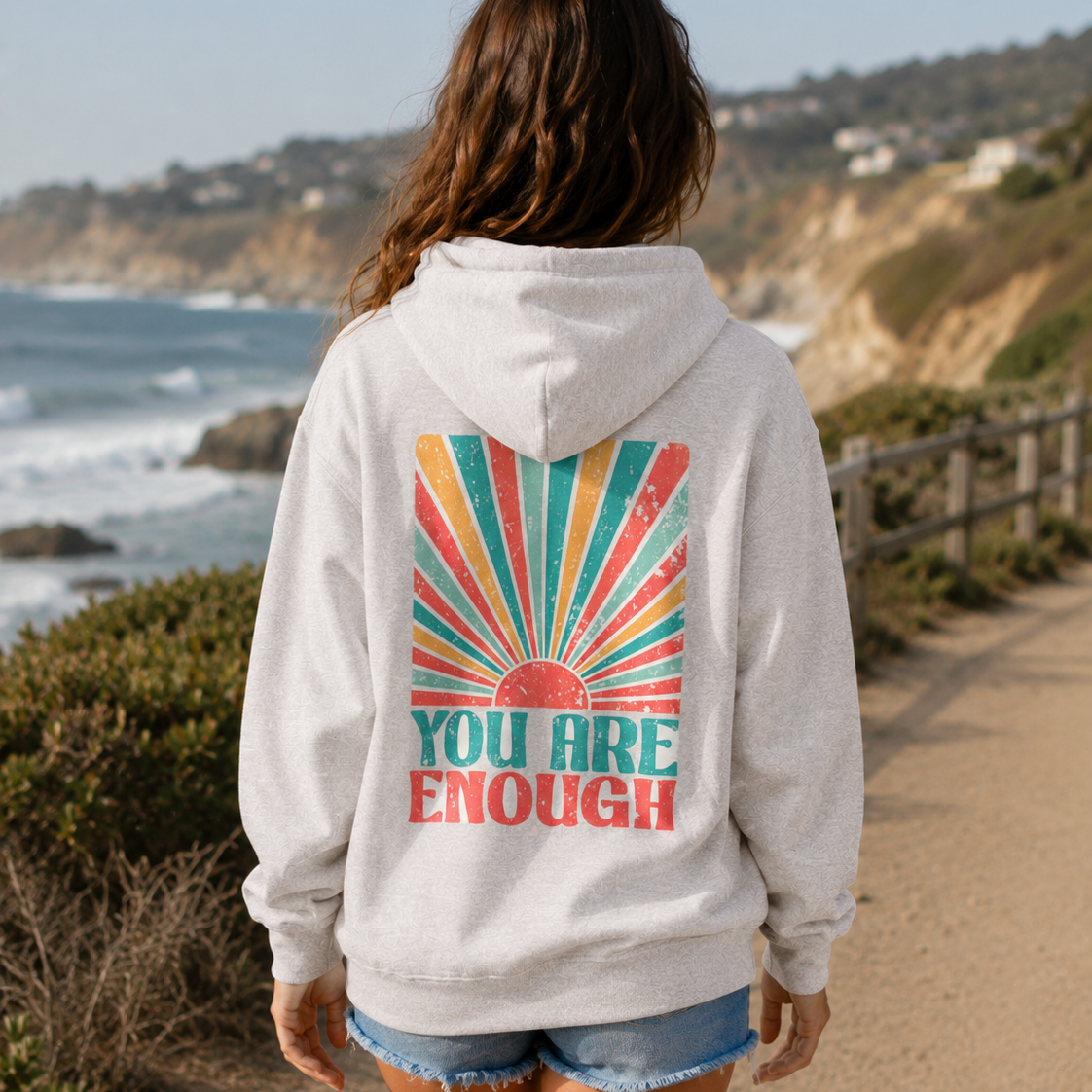 A woman viewed from behind wearing a light heather gray hoodie with a large retro-style back graphic that reads 'YOU ARE ENOUGH' in teal and coral over a multicolor sunburst. She is standing on a coastal path with ocean, cliffs, and hills in the background.