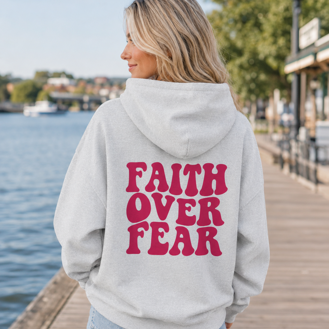 A woman standing on a wooden boardwalk by the water, wearing a light gray hoodie with the words 'FAITH OVER FEAR' printed in bold dark pink letters on the back, paired with light blue denim shorts.