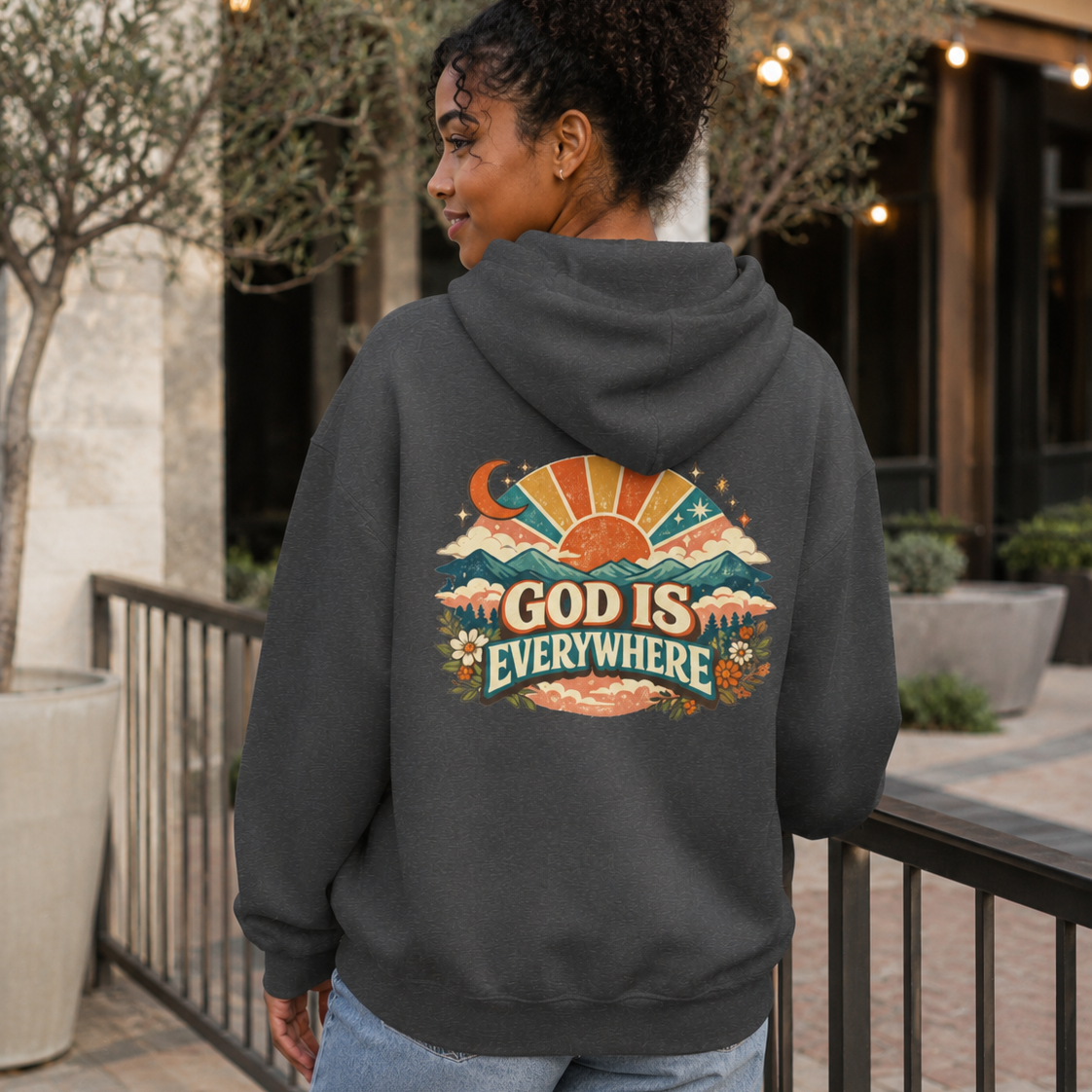 A woman wearing a dark gray hoodie with a colorful retro-style graphic on the back that reads 'GOD IS EVERYWHERE' with a sunset, mountains, and flowers, photographed in an outdoor patio setting.