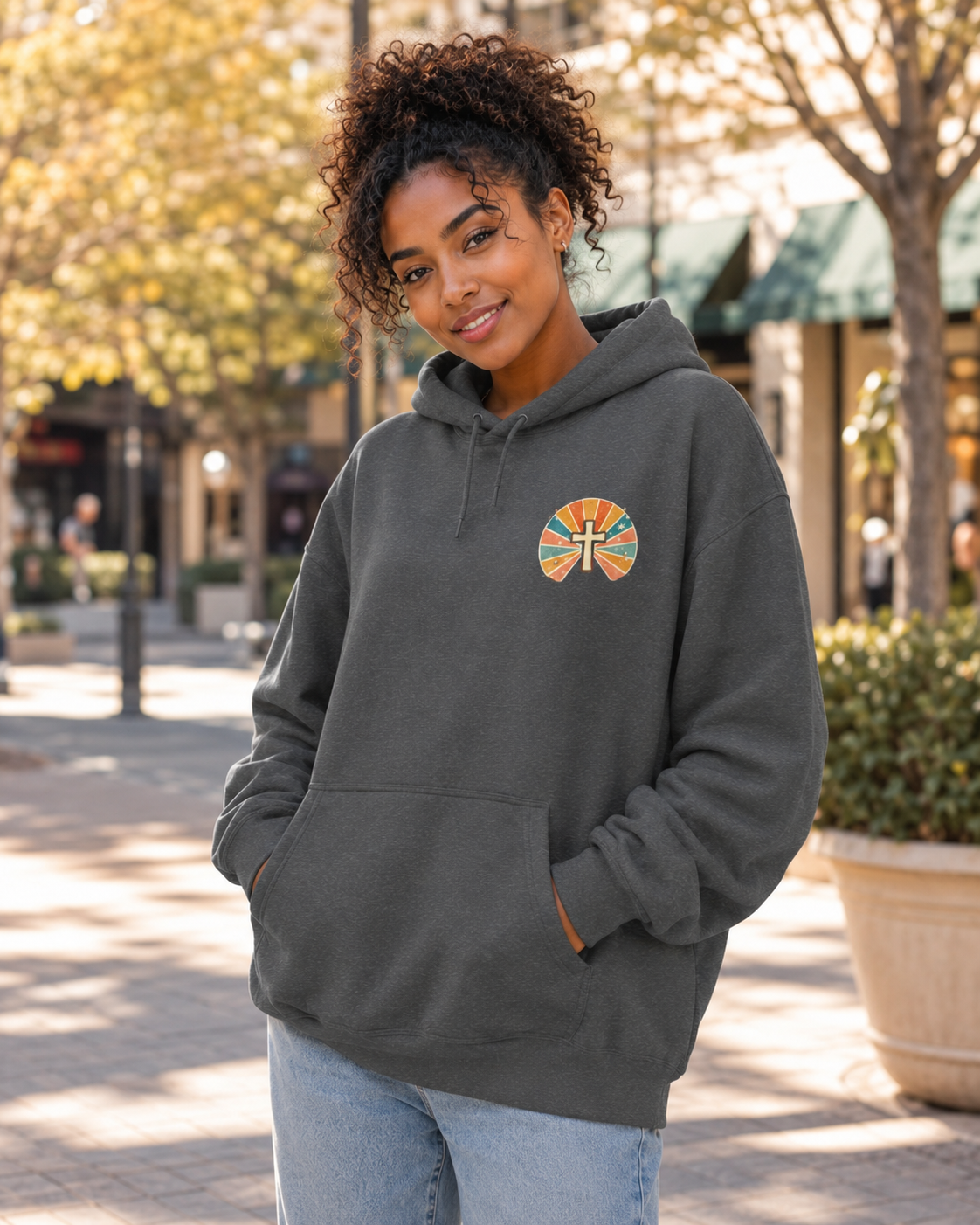A woman wearing a dark gray pullover hoodie with a colorful circular sunburst cross graphic on the left chest, standing outdoors on a paved walkway with trees and storefronts in the background.