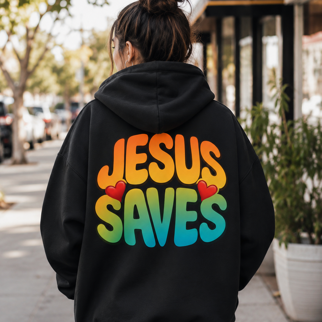 A person viewed from behind wearing a black hoodie with the phrase 'JESUS SAVES' printed across the back in a rainbow gradient, accented by two small red hearts. The person is walking on a sunny, tree-lined sidewalk.