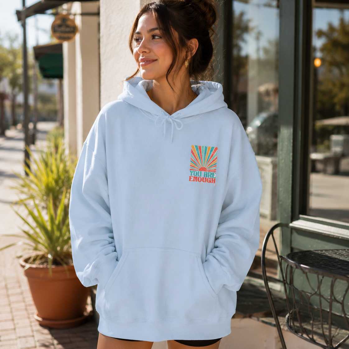 A woman wearing an oversized light blue hoodie with a colorful 'Sunshine California' graphic on the chest, standing outdoors in front of a storefront.