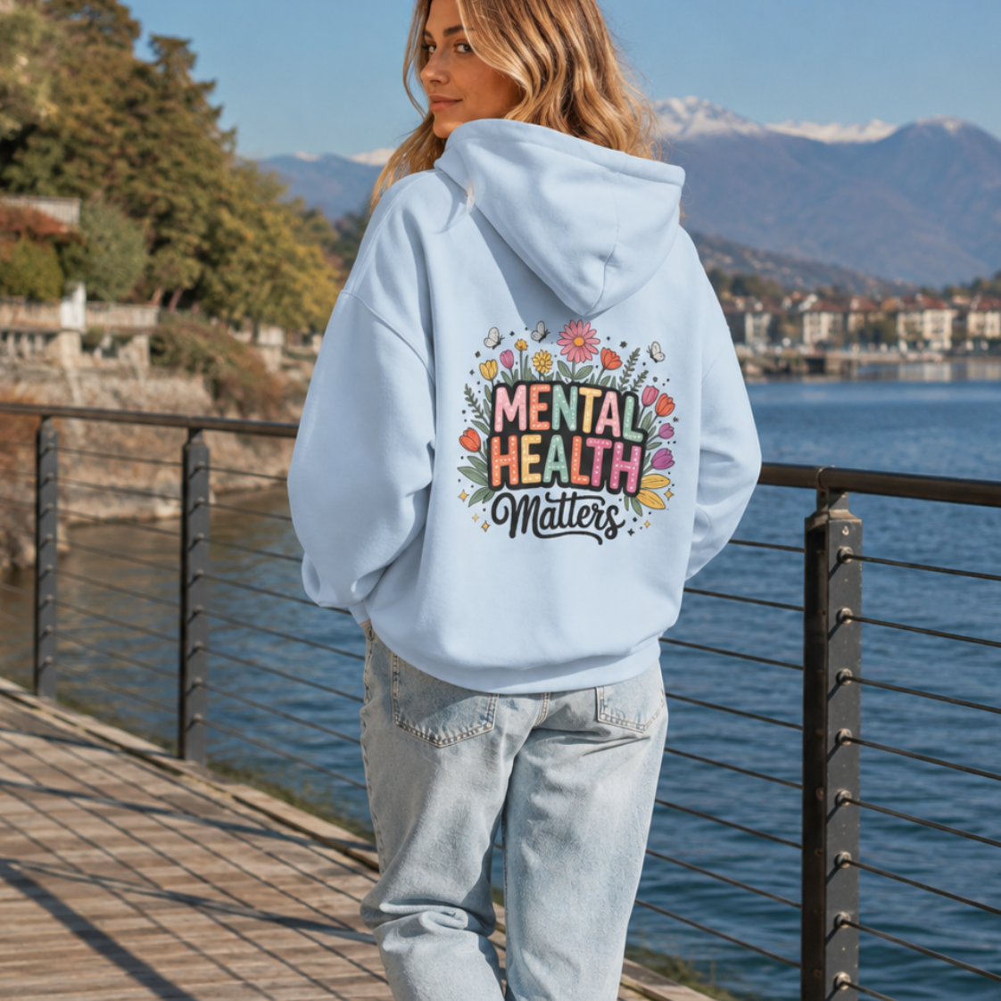 A woman walking on a wooden boardwalk by the water wearing a light blue hoodie with a colorful 'Mental Health Matters' graphic on the back, paired with light-wash jeans.