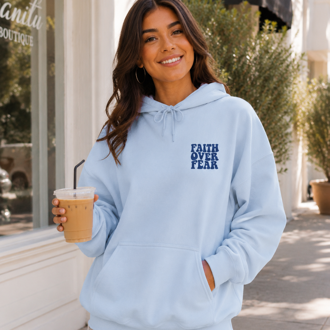 A woman wearing a light blue pullover hoodie with the text 'FAITH OVER FEAR' printed in dark blue on the left chest, holding an iced coffee on a sunny sidewalk.