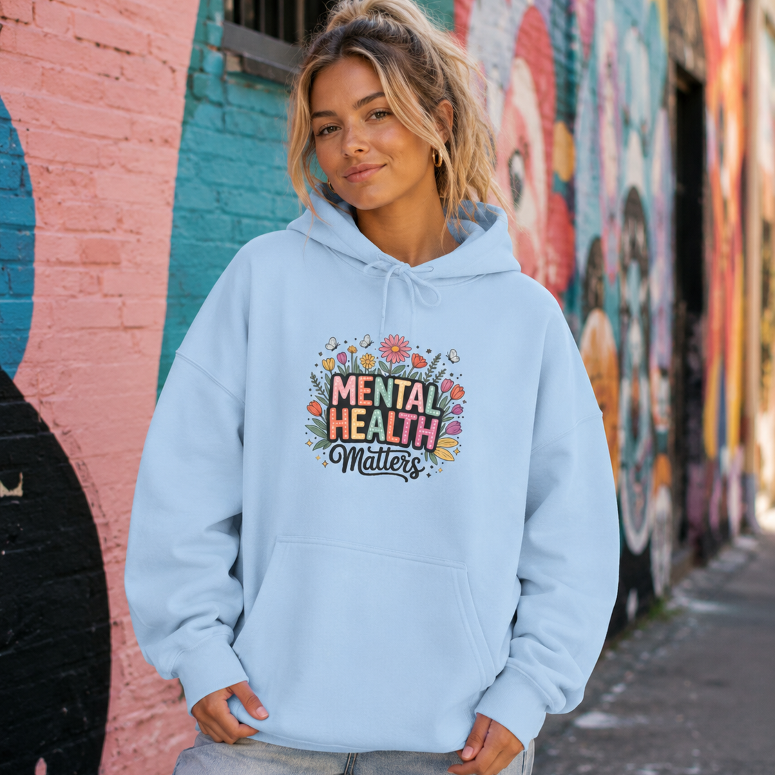 A woman wearing a light blue hoodie with a colorful 'Mental Health Matters' graphic, standing in front of a vibrant, painted brick wall.