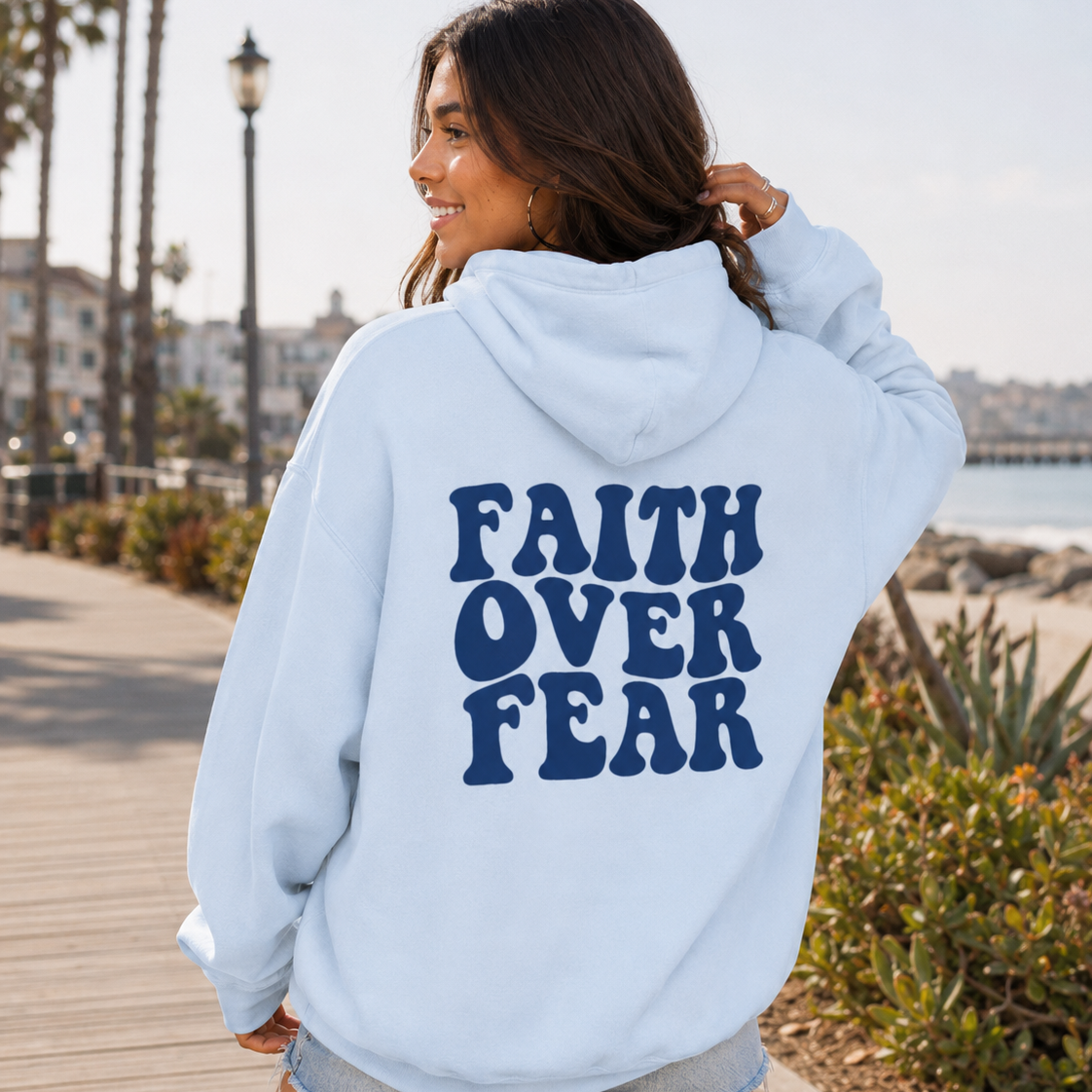 A woman wearing a light blue hoodie with the text 'FAITH OVER FEAR' printed in dark blue on the back, standing on a wooden boardwalk by the beach.