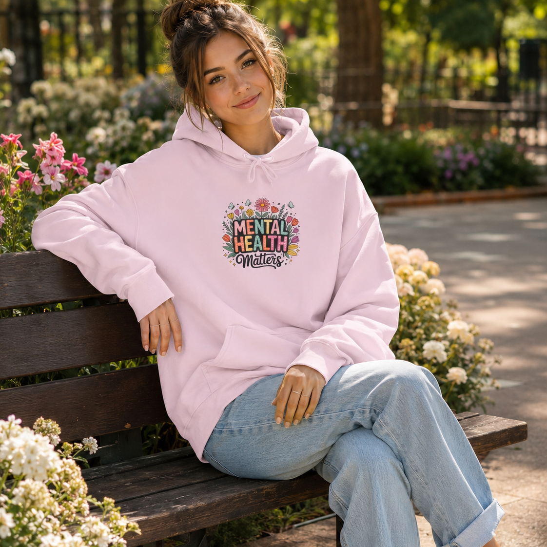 A woman sits on a wooden bench in a sunny park wearing a light pink hoodie with a colorful graphic that reads 'Mental Health Matters' and light blue jeans.