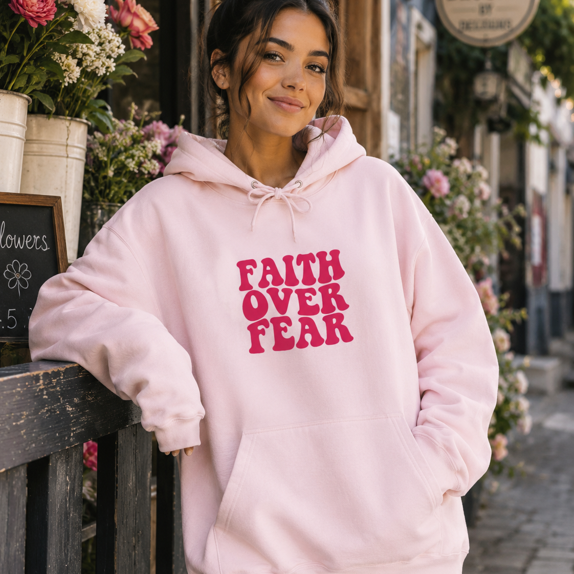 A woman wearing a light pink hoodie with the words 'FAITH OVER FEAR' printed in dark pink on the front, standing on a cobblestone street in front of a shop with flowers and a 'Blooms' sign.